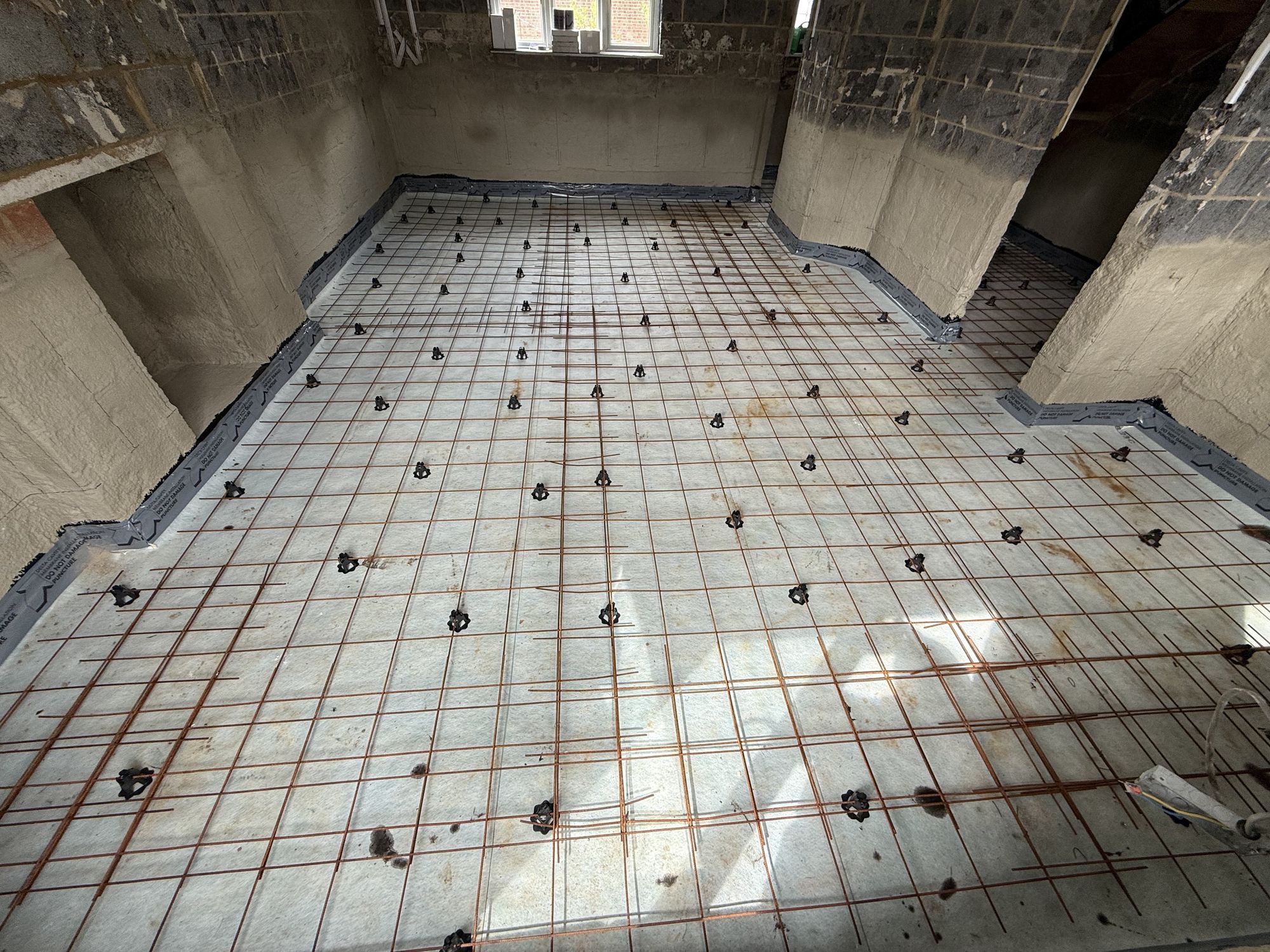 Felt-backed rubber waterproof tanking mat laid over the blinding sand, taped and sealed to the walls — steel mesh and chair spacers in position ready for the concrete slab pour