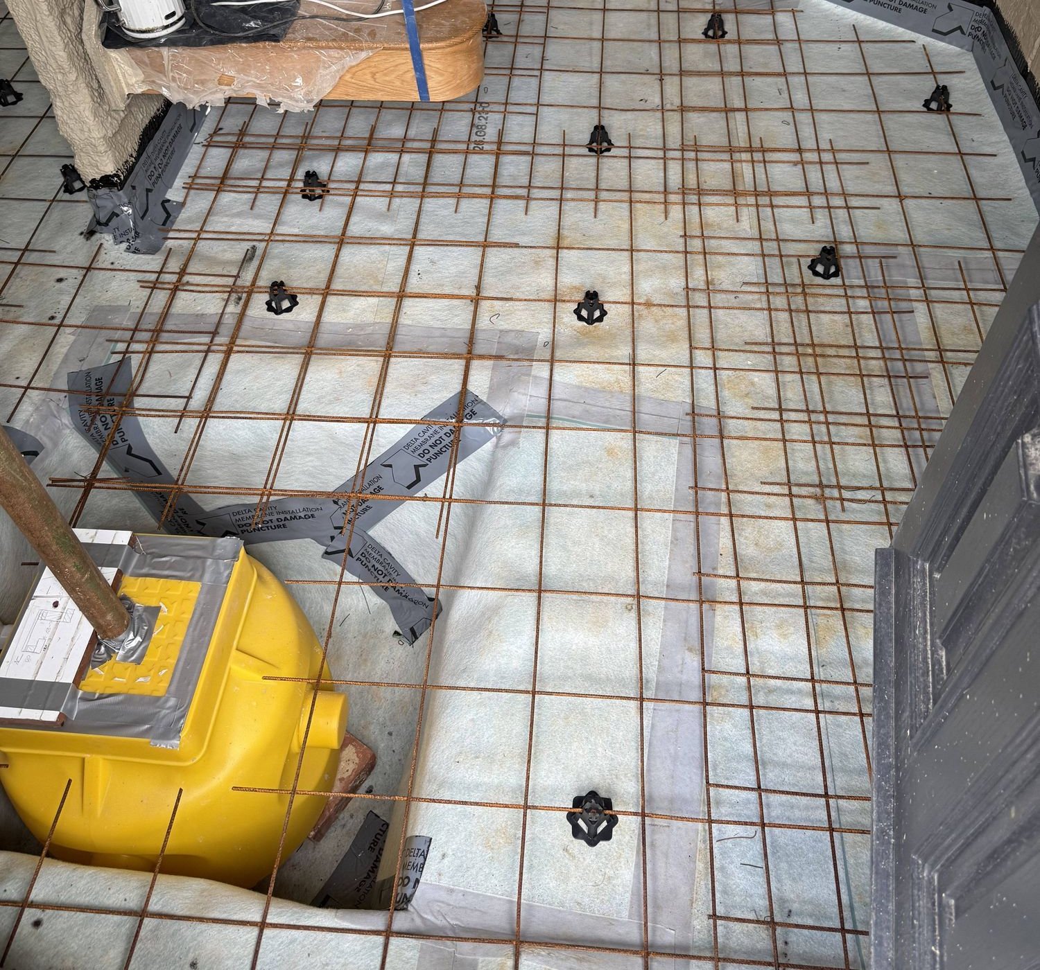 Felt-backed rubber waterproof tanking mat with steel mesh in position — yellow sump basket visible in the lower left corner